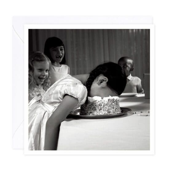 A black and white photo of a little girl dressed up in a beautiful white party dress with her head face down in a white frosted cake. Friends surround her.